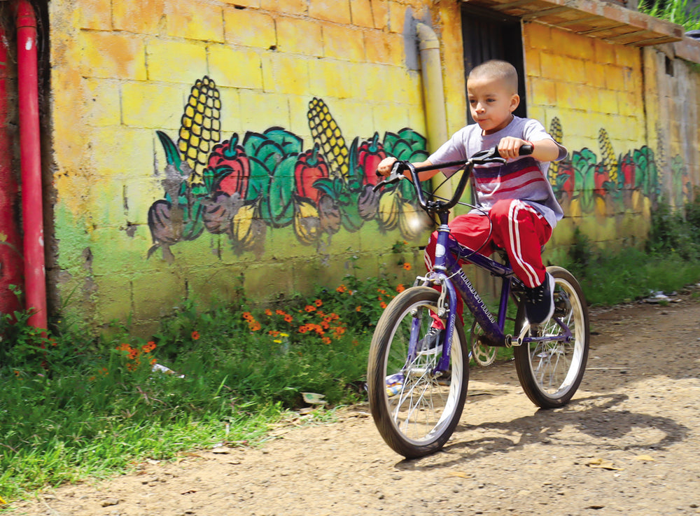 A young boy rides his bike down an alleyway, with a painted mural of vegetables behind him.
