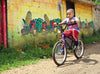 A young boy rides his bike down an alleyway, with a painted mural of vegetables behind him.