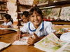 A young girl in a classroom smiles for the camera with a book in front of her.