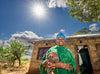 A woman smiles as she stands outside her home, holding a chicken under the bright blue sunlit sky.