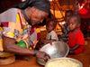 A grandmother serves food from a large bowl to her three grandchildren sitting on the ground, inside a tent-like structure.