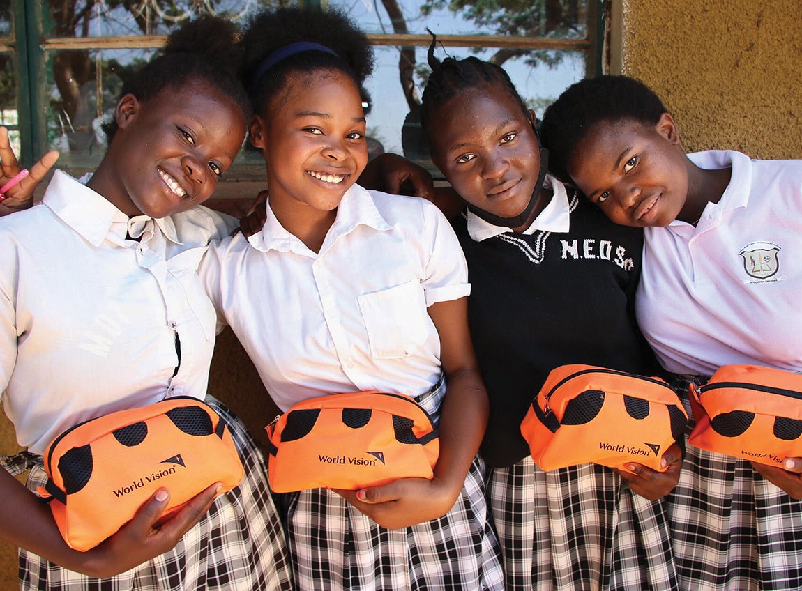 Four young girls in uniforms with plaid skirts smile for the camera. Each of them hold a small travel pouch that serves as a hygiene kit.