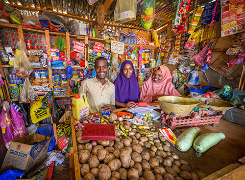 A man and two women smile as they stand inside the store they manage filled with various goods and produce.