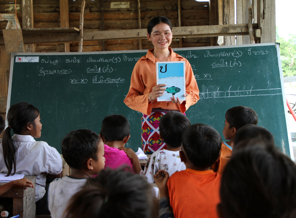 A woman stands in front of a chalkboard, holding a book towards children in the classroom.