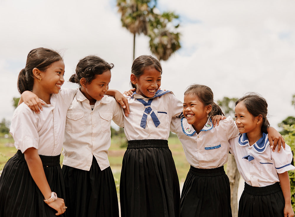 Five girls wearing blue and and white school uniform laugh and smile as they put their arms on 
each other's shoulders.