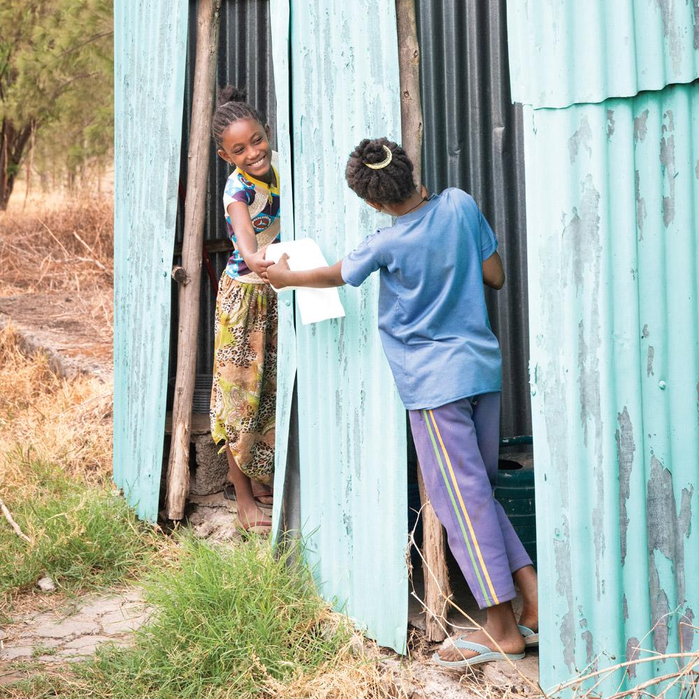Two girls standing at the entrance of latrines exchange toilet paper.