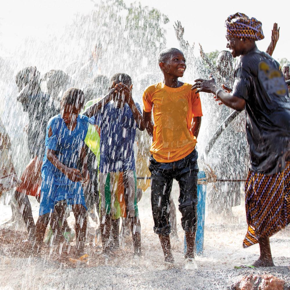 A group of children and an adult woman play in water coming out of a well.