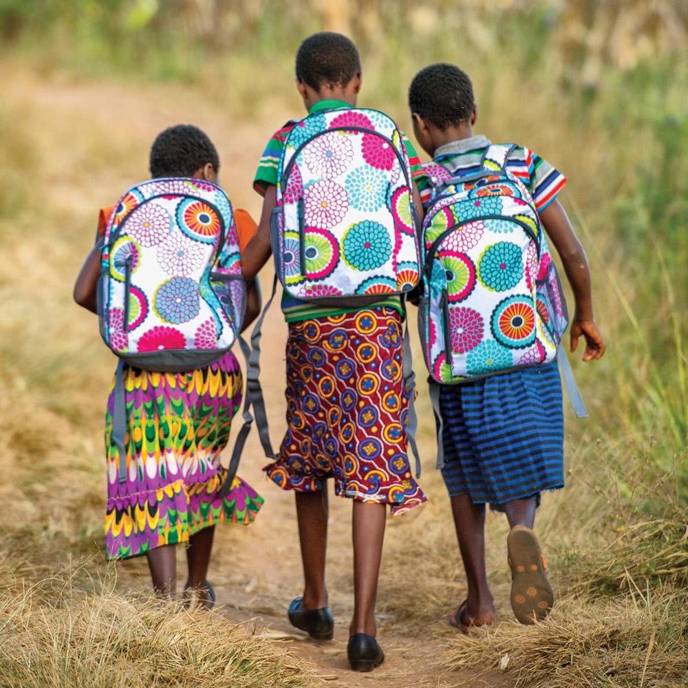 Three young girls carrying colorful backpacks walk side by side on a grassy area.