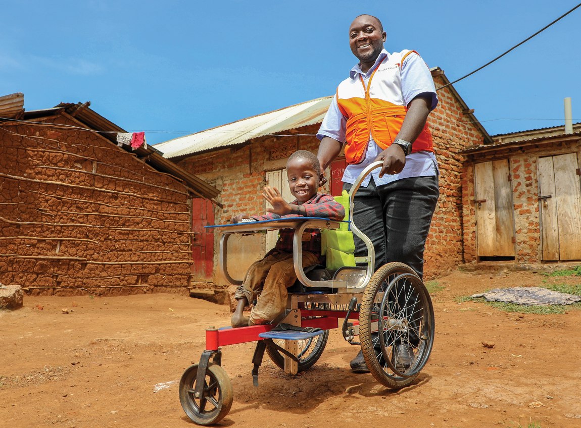 A smiling man wearing an orange and white World Vision vest pushes a smiling child in a wheelchair along a dirt road.