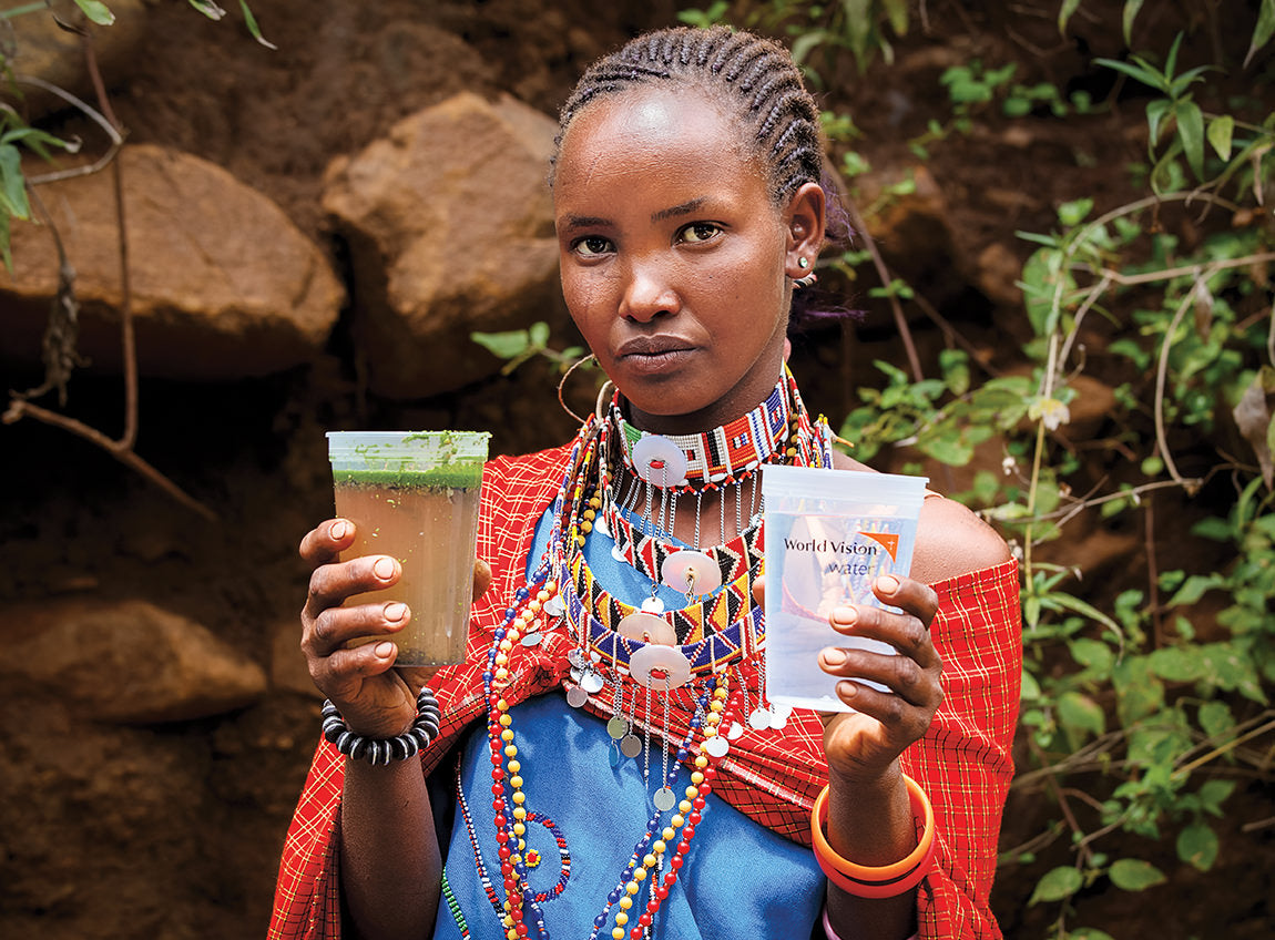 A woman wearing African clothing holds a cup of dirty water in her left hand and a cup of clean water in the other.